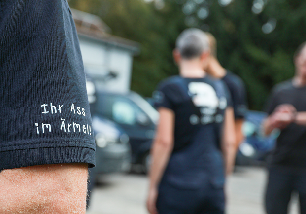 Nahaufnahme einer Person, die ein marineblaues Hemd mit der Aufschrift Ihr Arsch im Ärmel! trägt, während andere Personen unscharf im Hintergrund im Freien in der Nähe geparkter Autos stehen.