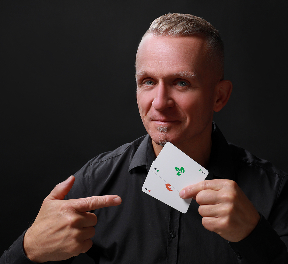 Ein Mann in einem schwarzen Hemd lächelt und zeigt auf eine Spielkarte, die er in der Hand hält. Die Karte zeigt ungewöhnliche grüne und rote Symbole anstelle der traditionellen Farben. Der Hintergrund ist einfarbig schwarz.