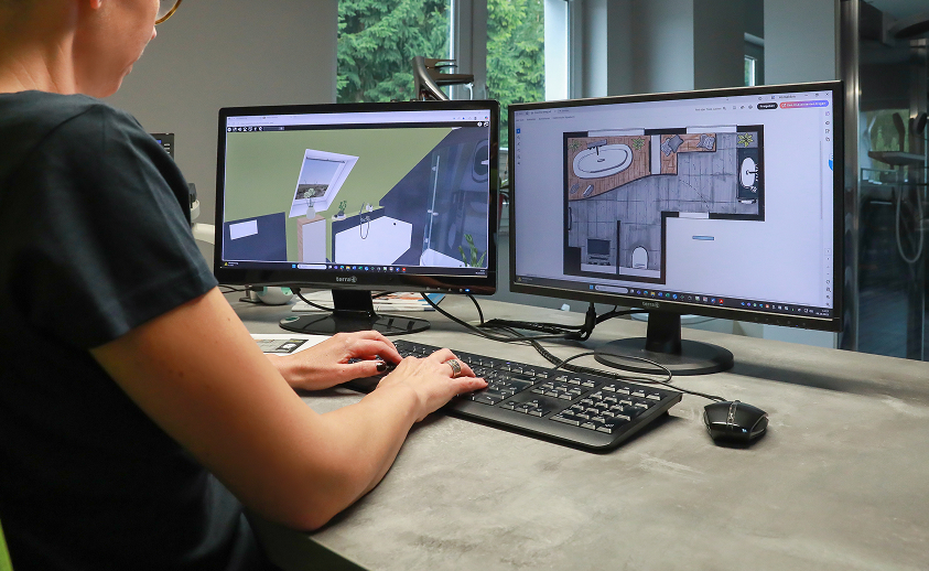 Eine Person arbeitet an einem Schreibtisch mit zwei Computermonitoren, auf denen eine Software für Architektur oder Innenarchitektur läuft, die auf einem Bildschirm ein 3D-Raummodell und auf dem anderen einen Grundriss zeigt.