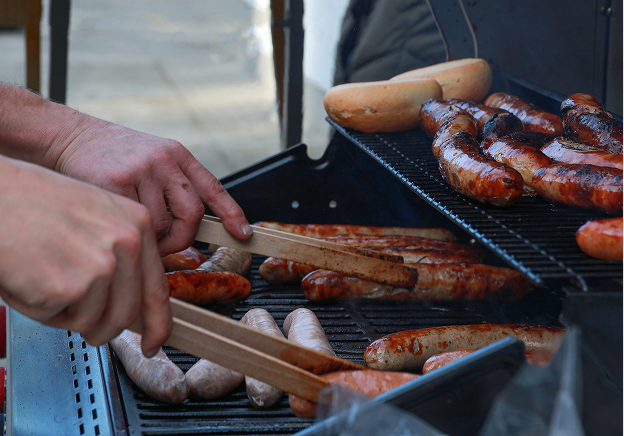 Eine Person benutzt eine Zange, um Würstchen und Bratwürste auf einem Grill zu grillen, wobei die Brötchen auf dem oberen Rost geröstet werden. Die Bratwürste brutzeln und sind gebräunt, so dass eine lebhafte Kochszene im Freien entsteht.