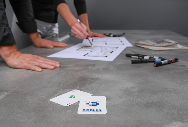 Zwei Personen arbeiten an einem grauen Tisch mit Markern und Papieren an Architekturplänen, während im Vordergrund zwei Spielkarten zu sehen sind, darunter ein Kreuz-Ass und eine Markenkarte. Zwei Personen arbeiten an einem grauen Tisch mit Markern und Papieren an Architekturplänen, während im Vordergrund zwei Spielkarten zu sehen sind, darunter ein Kreuz-Ass und eine Markenkarte.