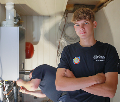 Ein junger Mann in einem schwarzen T-Shirt steht mit verschränkten Armen da und lächelt in die Kamera, während eine andere Person im Hintergrund in einer Waschküche an Rohren arbeitet. Ein junger Mann in einem schwarzen T-Shirt steht mit verschränkten Armen da und lächelt in die Kamera, während eine andere Person im Hintergrund in einer Waschküche an Rohren arbeitet.