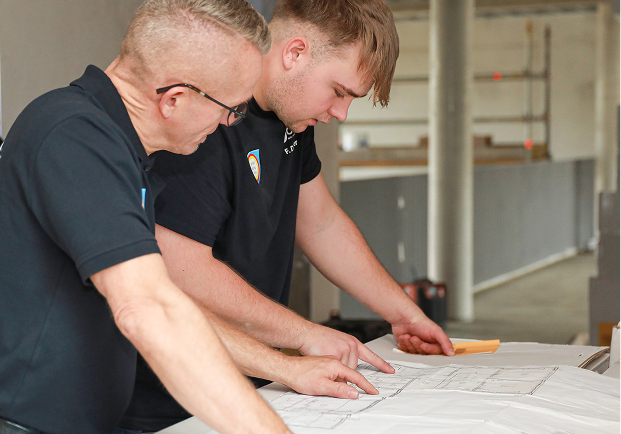 Zwei Männer in schwarzen Hemden begutachten und diskutieren Architekturpläne auf einem Tisch in einem hellen, unfertigen Innenraum.