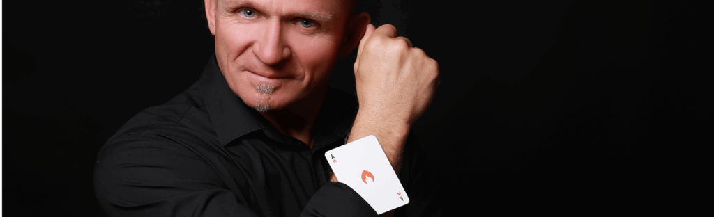 Start 2 Ein Mann in einem schwarzen Hemd lächelt leicht, während er seinen rechten Arm hochhält und eine Pik-As-Karte unter seinem Ärmel vor einem schwarzen Hintergrund zeigt.