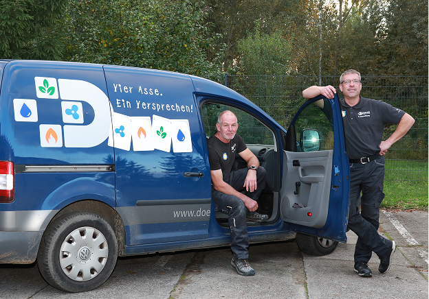 Zwei Männer in Arbeitsuniformen posieren vor einem blauen Lieferwagen. Einer sitzt in der offenen Fahrertür, während der andere an der Tür lehnt. Auf der Seite des Lieferwagens sind bunte Symbole und deutsche Texte zu sehen. Im Hintergrund sind Bäume und Gras zu sehen.