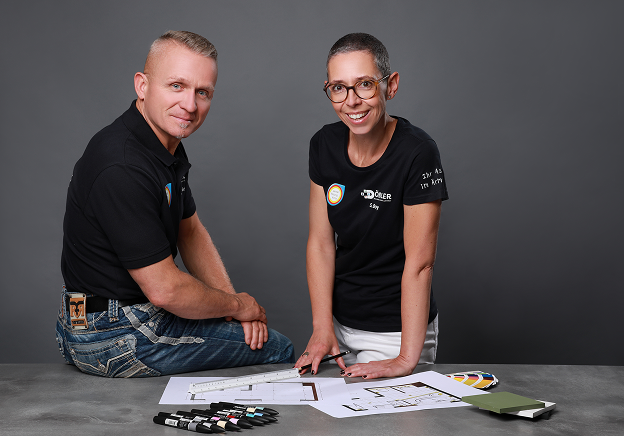 Zwei Personen, ein Mann und eine Frau, lächeln in die Kamera, während sie sich über einen Tisch mit Bauplänen, Markern, Farbmustern und Papier beugen. Sie sind leger gekleidet und stehen vor einem schlichten grauen Hintergrund.
