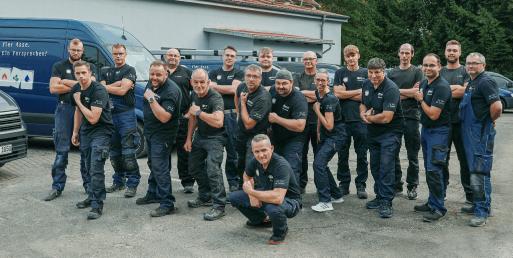 Eine Gruppe von sechzehn Männern in passenden dunklen Hemden und Arbeitshosen posiert selbstbewusst mit ausgestreckten Armen vor einem blauen Firmenwagen im Freien. Die meisten lächeln und stehen auf einer gepflasterten Fläche, hinter der sich Bäume und ein Gebäude befinden.