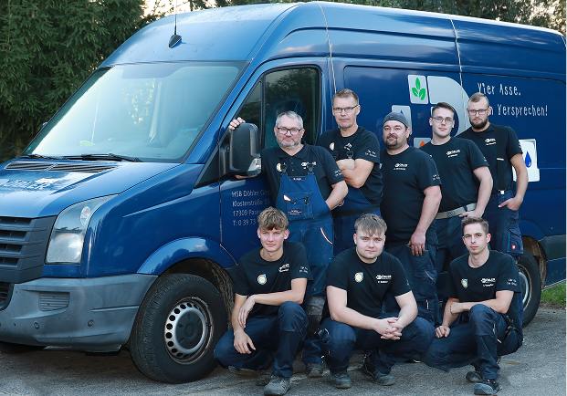 Acht Männer in passenden dunklen Hemden posieren gemeinsam neben einem blauen Arbeitswagen. Einige stehen, andere kauern vor dem Lieferwagen, und sie alle scheinen Teil eines Teams oder einer Mannschaft zu sein. Im Hintergrund sind Bäume zu sehen.