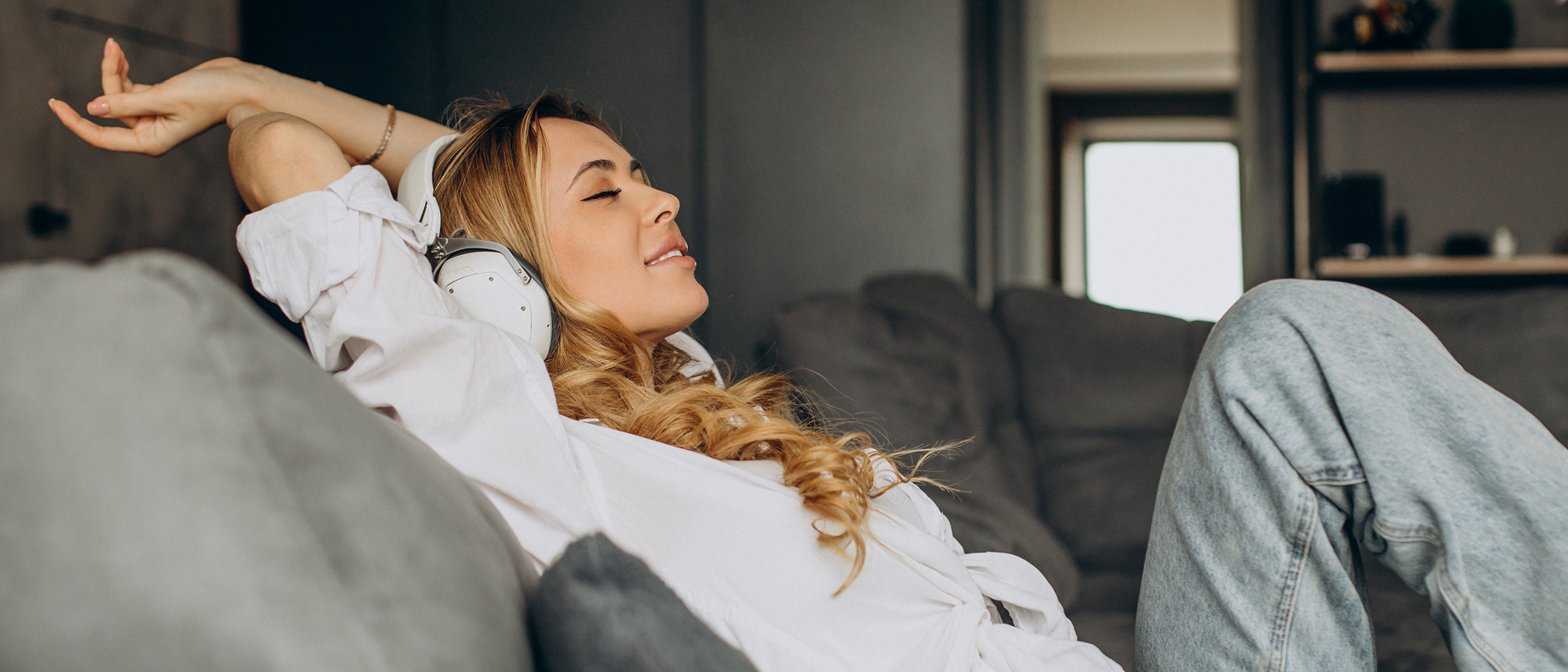 Eine junge Frau mit langen Haaren entspannt sich auf einer grauen Couch, trägt Kopfhörer und ein weißes Hemd mit blauen Jeans. Sie lehnt sich mit geschlossenen Augen und einem über den Kopf gestreckten Arm zurück und genießt die Musik.