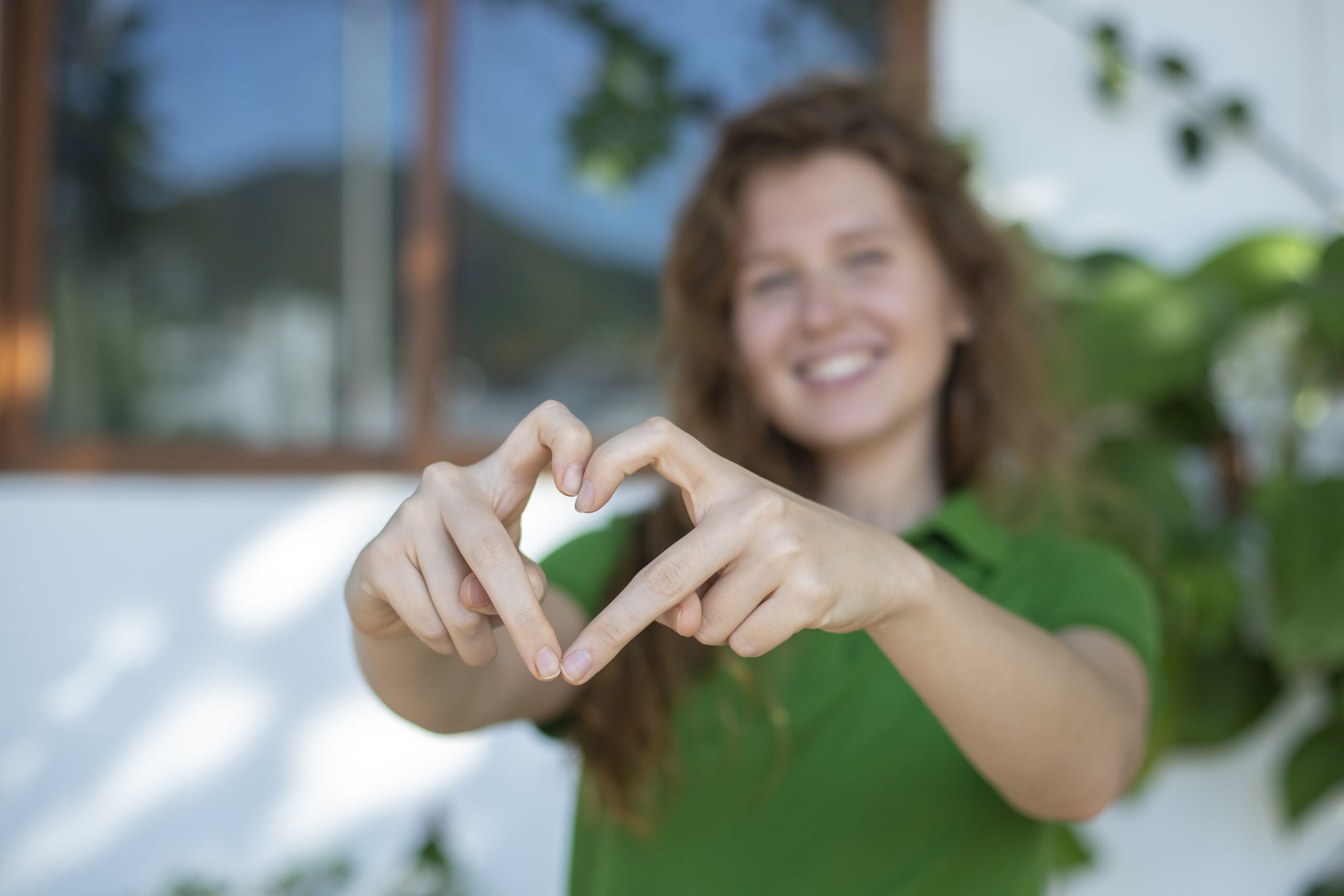 Eine Person mit langem, gewelltem Haar und grünem Hemd steht im Freien, lächelt und formt mit ihren Händen ein Herz, vor einem unscharfen Hintergrund aus Grün und einem Fenster.