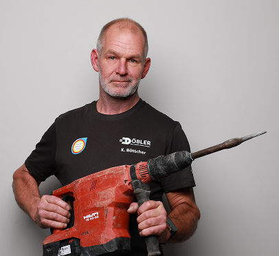 Ein bärtiger Mann mittleren Alters, der ein schwarzes Hemd mit Aufschrift trägt, hält eine große rote Bohrmaschine mit einem langen Bohrer vor einem schlichten hellgrauen Hintergrund.