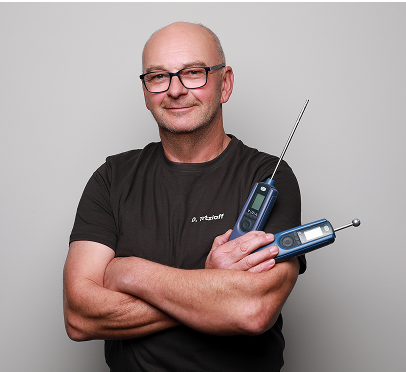 Ein lächelnder Mann mit Brille steht mit verschränkten Armen vor einem schlichten Hintergrund und hält zwei digitale Thermometer in der Hand - eines mit einer Metallsonde und das andere mit einem runden Sensor.